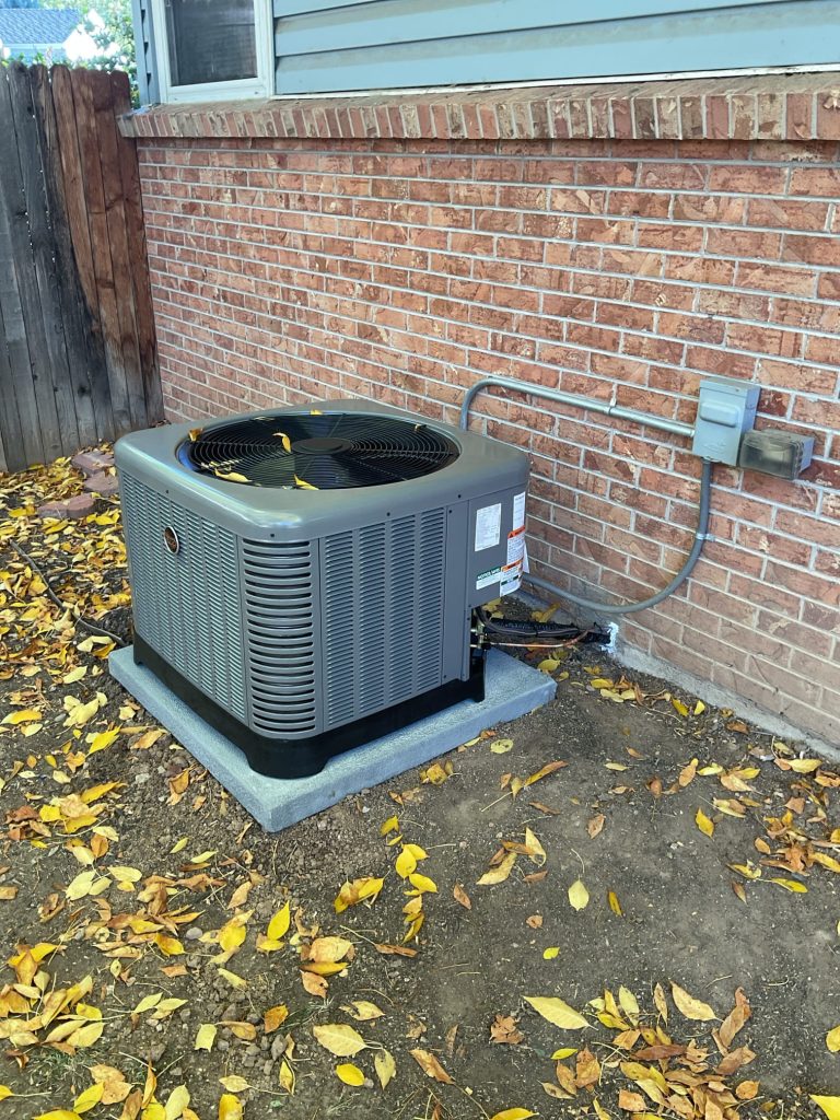 A central air conditioning unit is installed on a concrete pad next to a brick house, with electrical wiring connected and fallen yellow leaves scattered on the ground.