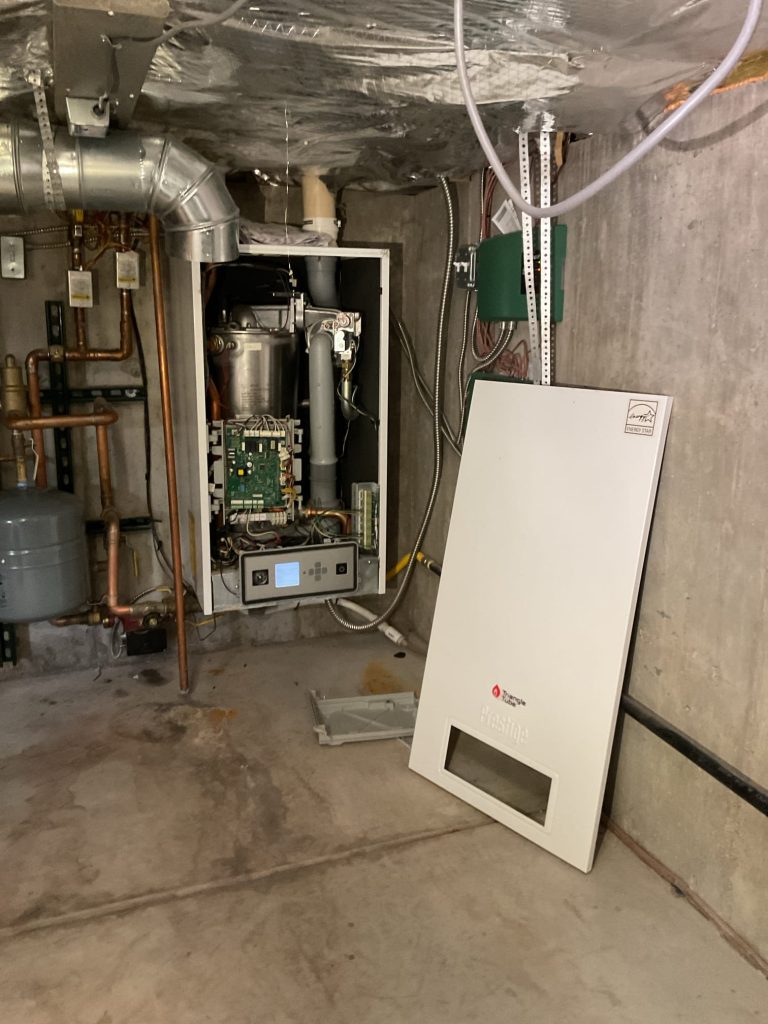 An open boiler unit with its front panel removed sits in a basement with exposed pipes and concrete walls; the panel is leaning against the wall on the floor. An open boiler unit with its front panel removed sits in a basement with exposed pipes and concrete walls; the panel is leaning against the wall on the floor.