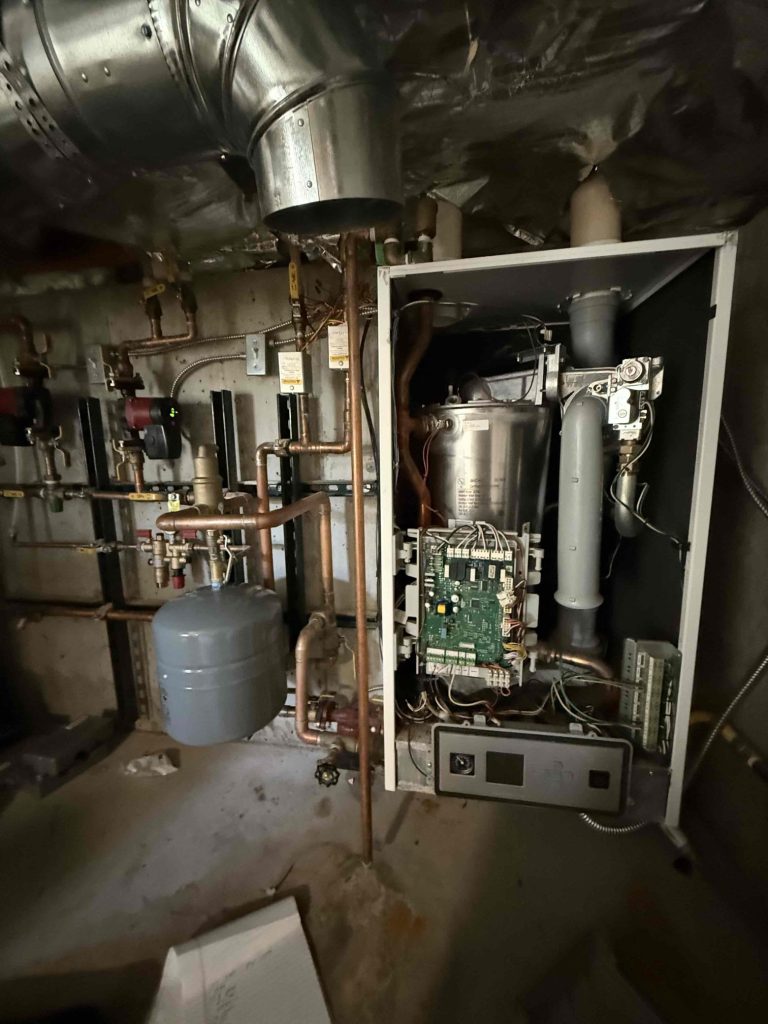 An open boiler unit with visible internal components and circuit board, surrounded by various pipes, valves, and a pressure tank in a dimly lit utility room. An open boiler unit with visible internal components and circuit board, surrounded by various pipes, valves, and a pressure tank in a dimly lit utility room.